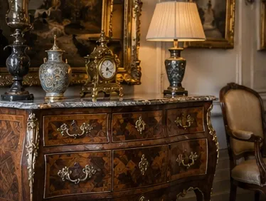 Commode galbée en marqueterie à poignées de bronze doré et plateau de marbre, surmontée d'une pendule et de vases, avec un fauteuil ancien et des tableaux dorés
