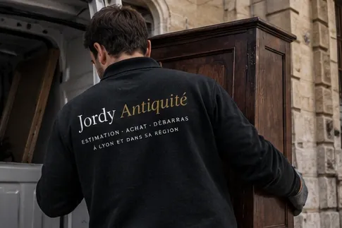 Un homme vu de dos, en sweat Jordy Dellinger, déplace une armoire ancienne en bois devant une camionnette